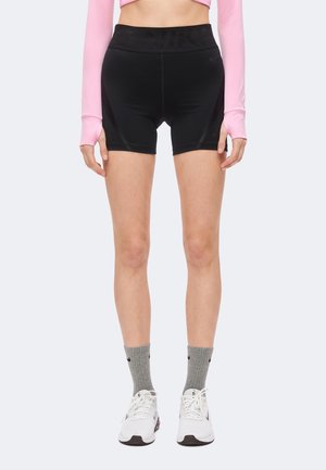 Femme portant un short de sport noir taille haute, un crop top rose, des chaussettes grises et des baskets blanches, debout devant un fond uni.