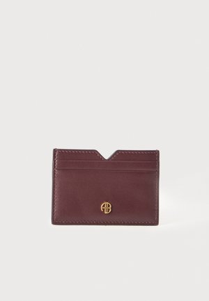 Porte-cartes en cuir bordeaux aux bords surpiqués, avec deux fentes pour cartes, une ouverture supérieure échancrée et un petit monogramme doré, sur fond clair.