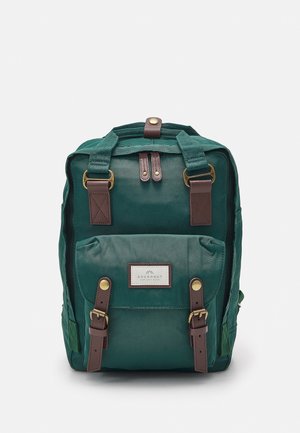 Sac à dos en toile verte avec poche avant, bretelles en cuir marron, boucles en laiton et étiquette indiquant "Doughnut" sur fond blanc.