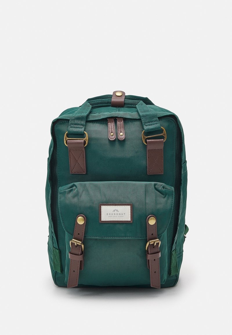 Sac à dos en toile verte avec poche avant, bretelles en cuir marron, boucles en laiton et étiquette indiquant "Doughnut" sur fond blanc.