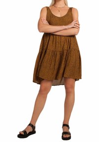 Mujer de pie con los brazos cruzados, vistiendo un vestido sin mangas de estampado marrón y sandalias negras con tiras al tobillo, sobre un fondo blanco.