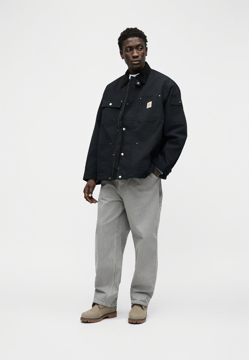 Man wearing a black work jacket with front pockets, striped loose pants, and beige boots, standing against a plain white background.