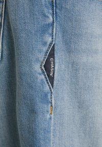 Tela de mezclilla azul clara con una superficie texturizada. Presenta un bolsillo en la costura lateral con una etiqueta azul oscura que dice "CHASIN'" cosida en él.