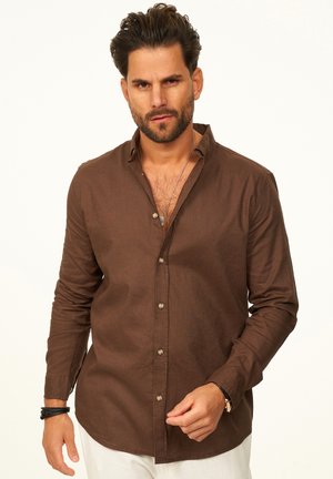 Mann mit gestyltem dunklem Haar und Bart, der ein braunes Hemd mit Knöpfen, weiße Hose und schwarze Armbänder trägt, vor einem hellen Hintergrund steht.