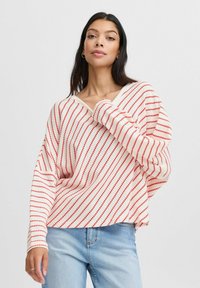 Pull en tricot rayé rouge et blanc avec un décolleté en V, coupe ample et poignets côtelés, présentant un tissu texturé et un motif de rayures diagonales.