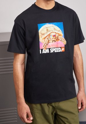 Personne portant un t-shirt noir avec un graphique d'une tortue avec des lunettes de soleil et le texte "I AM SPEED" au-dessus d'un logo Nike rouge.