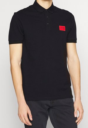 Mann trägt ein schwarzes Polohemd mit kurzen Ärmeln, drei Knöpfen und kleinem roten "HUGO"-Logo auf der linken Brust.