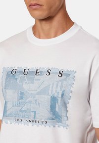 Biała bawełniana koszulka z niebieskim nadrukiem przedstawiającym panoramę miasta z napisem „GUESS” i „LOS ANGELES”, okrągły dekolt i krótkie rękawy.