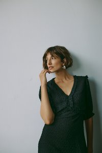 Crna haljina od teksturirane tkanine, s volanima, v-izrezom i kratkim rukavima. Model pozira u odnosu na jednostavnu zidnu pozadinu.