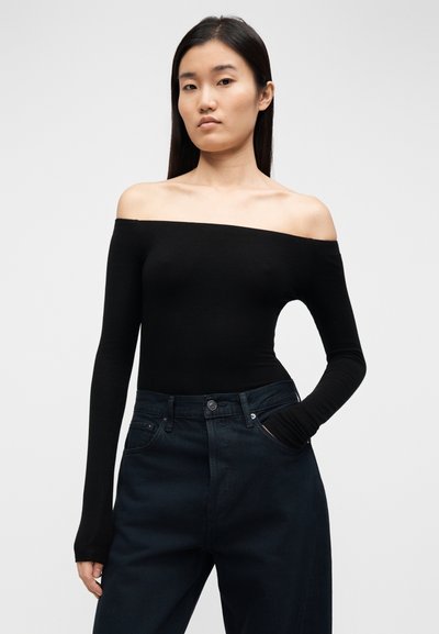 Čierny top s dlhým rukávom so zospodu odhalenými ramenami, priliehavý strih, mäkká látka, nosený s tmavými, vysoko vystrihnutými džínsami s predným vreckom.
