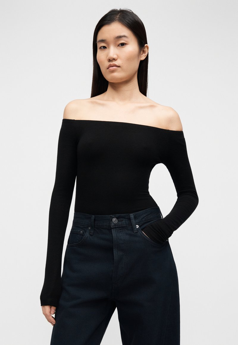 Haut noir à manches longues, épaules dénudées, coupe ajustée, tissu doux, porté avec un jean foncé taille haute avec une poche avant.