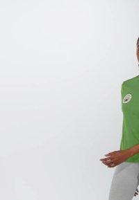 Green cotton t-shirt with a round neckline and short sleeves, featuring a round patch on the left side. Worn with light gray pants.
