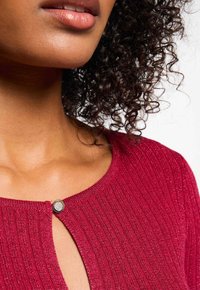 Maglione rossa lavorato a maglia con texture a costine, scollatura rotonda e un bottone decorativo con una gemma accanto alla patta.