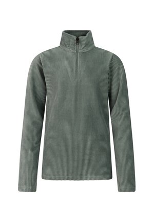Junge Frau trägt eine schwarze Mütze, grünes geripptes Langarmshirt, schwarze Hose und schwarze Schuhe, steht vor einem schlichten Hintergrund.
