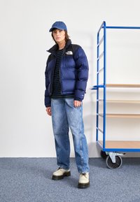 Marine pufferjakke med sort krave, stribet skjorte, lys blå jeans og off-white platformsko. Blå kasket og bagagevogn i baggrunden.