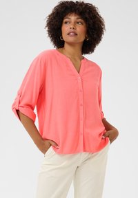 Camicia corallo con bottoni, col V e maniche arrotolate. Tessuto leggero con una texture liscia. Indossata con pantaloni chiari.