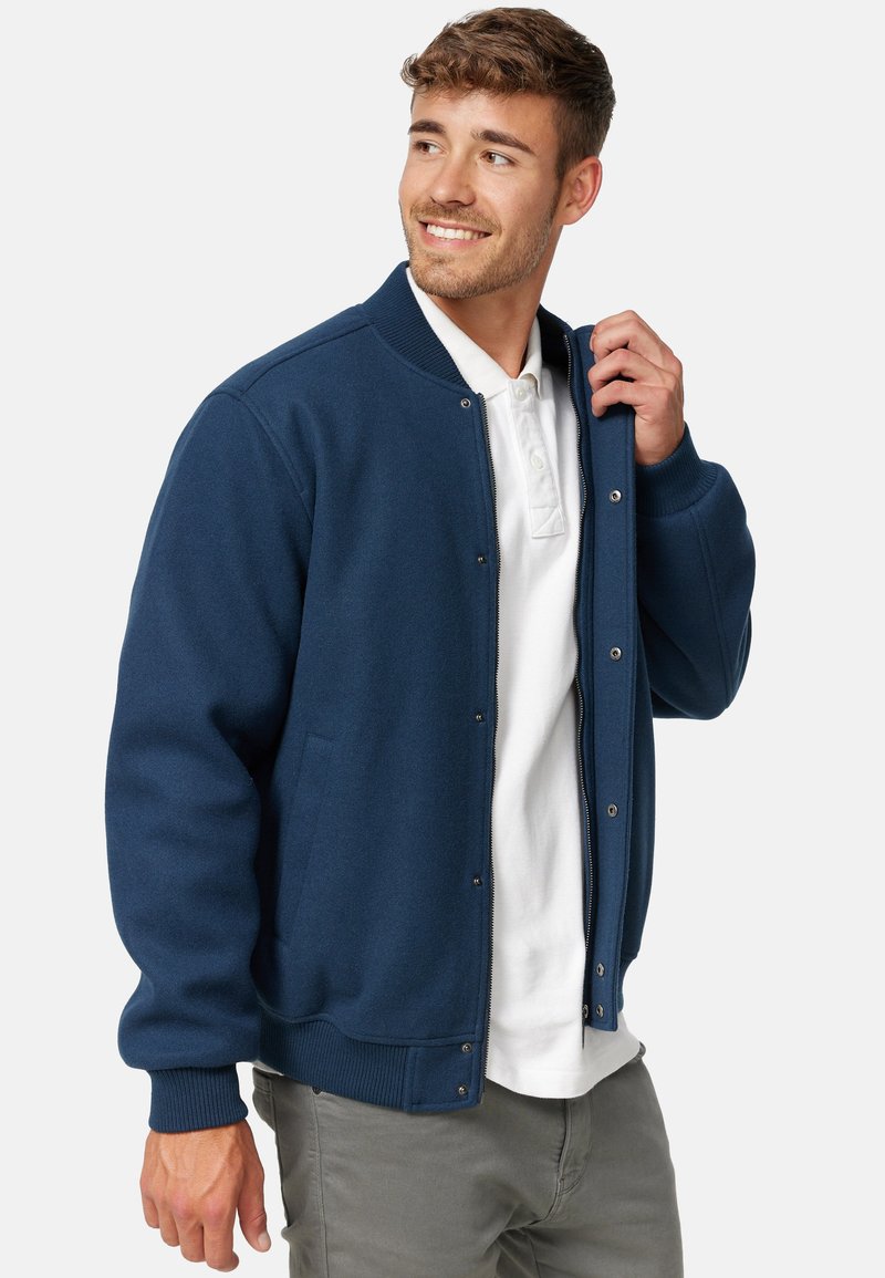 Marineblauwe bomberjack met een rits aan de voorkant, ribbelmanchetten en -zoom, twee zijzakken en een zachte textuur. G gedragen over een wit poloshirt.