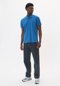 Polo de manga corta en tejido de algodón azul brillante, con un cuello clásico y una tapeta de tres botones, combinado con jeans de mezclilla oscura y zapatillas deportivas.