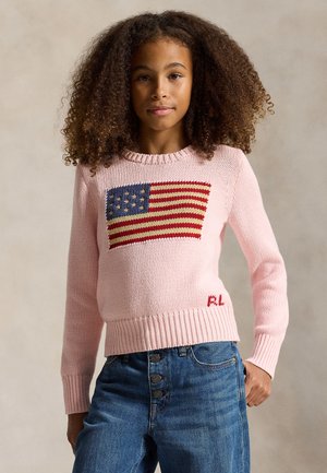 Jeune fille aux cheveux bouclés portant un pull rose avec un motif du drapeau américain et un jean bleu, debout devant un fond neutre.