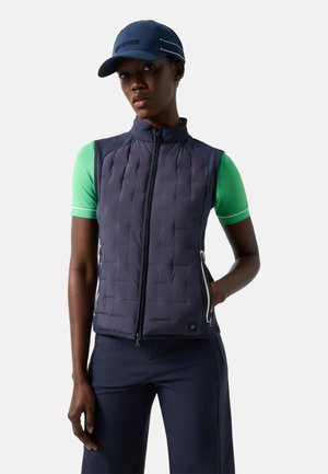 Frau trägt eine marineblaue gesteppte Weste über einem grünen kurzärmligen Shirt, marineblaue Hose und marineblaue Kappe, steht mit einer Hand in der Tasche.