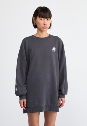 Una joven con cabello oscuro lacio lleva un vestido sudadera gris oscuro de gran tamaño con estampados de sol y estrellas en el pecho y la manga.