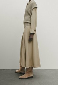 Beige strikket sweater vest over en beige langærmet skjorte og en let, plisseret nederdel, parret med lysebrune ruskindsankelhøje og en glat finish.