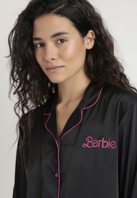 Camisa de pijama de satén negro con ribete rosa y el logotipo bordado de "Barbie", que presenta un cuello clásico y un detalle de cierre de botón.