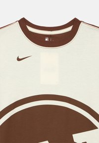 Brązowo-kremowy T-shirt z krótkim rękawem i okrągłym dekoltem, z dużym okrągłym logo oraz małym znakiem Nike Swoosh na piersi.