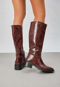 Bottes bordeaux jusqu'au genou avec une texture croco embossée brillante et un talon noir empilé. Comprend une fermeture éclair sur le côté pour un enfilage facile.