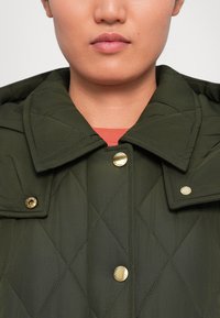 Prim-plan cu o persoană îmbrăcată într-o jachetă matlasată de culoare verde închis, cu nasturi aurii și un guler mare, purtată peste un top roșu.