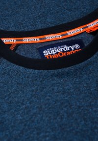 Close-up van een blauwe, gestructureerde Superdry The Orange sweatshirtkraag met zwarte rand en een gevoerde oranje branding aan de binnenkant.