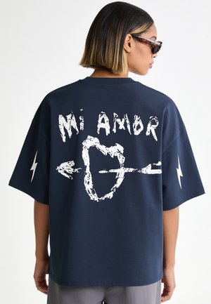 Donna che indossa una t-shirt oversize blu scuro con testo ""Mi Amor"" e grafica di cuore trafitto da una freccia sulla schiena.