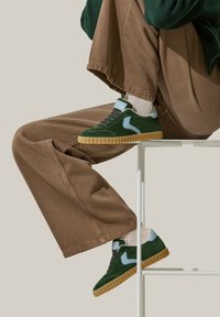 Baskets en daim vert avec des accents bleu clair, semelle en caoutchouc texturée, lacets foncés et minimalisme au niveau du branding, portées avec un pantalon en velours côtelé marron.