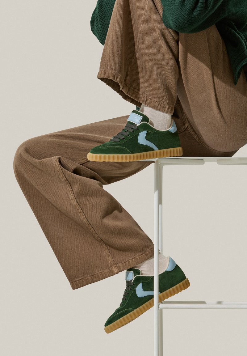 Baskets en daim vert avec des accents bleu clair, semelle en caoutchouc texturée, lacets foncés et minimalisme au niveau du branding, portées avec un pantalon en velours côtelé marron.
