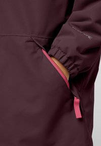Bordeauxfarbene wasserdichte Jacke mit elastischen Bündchen, sichtbarem pinken Reißverschluss und seitlichen Taschen. Glatte Textur, elegantes Design und minimale Markenkennzeichnung.