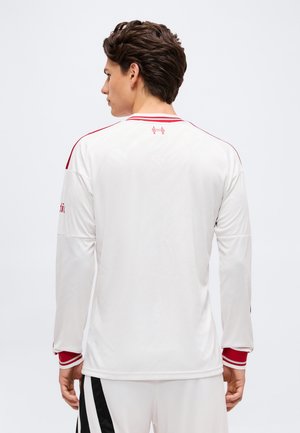 Jeune homme aux cheveux bruns courts portant un maillot de sport blanc à manches longues avec des bordures rouges, vu de dos sur un fond uni.