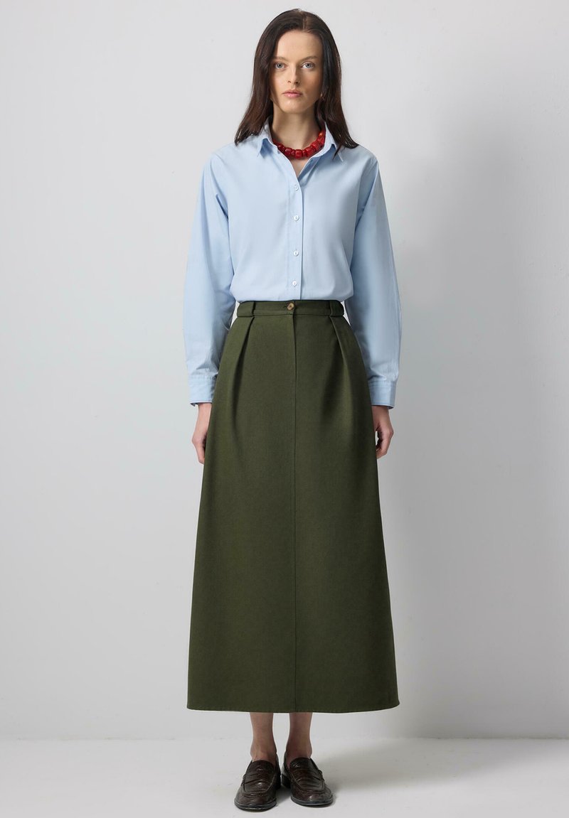 Γυναίκα που στέκεται φορώντας φωτεινό γαλάζιο πουκάμισο με κουμπιά, μακριά φούστα ψηλόμεση σε ελιά πράσινο, κόκκινο κολιέ με χάντρες και σκούρα καφέ μοκασίνια.