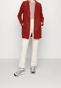 Cardigan rojo de manga larga con bolsillos, usado sobre una blusa rosa clara, combinado con pantalones de color crema y zapatillas plateadas. Diseño simple.