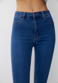 Jeans de tiro alto en azul, fabricados en denim, con una textura suave, bolsillos frontales visibles y detalles de costura en color naranja.