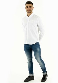 Chemise blanche à boutons avec col, associée à un jean bleu ajusté et des baskets noires. La chemise présente un petit logo vert sur la poitrine.