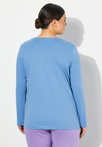 Dlouhý rukáv top v světle modré bavlně s zaobleným vpředu a rovnými okraji; spárovaný s fialovými kalhotami. Hladká textura, minimální švy.