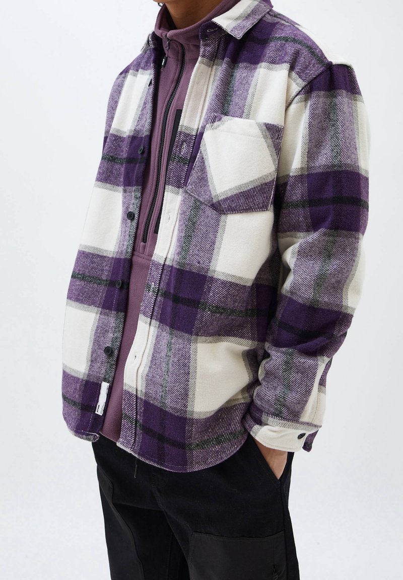 Personne portant une chemise à carreaux violette et blanche boutonnée par-dessus un sweat zippé violet avec un pantalon noir, debout avec une main dans la poche.