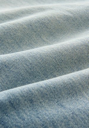Ljusblå denimtyg med fina vertikala ribbningar, som visar en mjuk textur och subtila färgvariationer över vikta ytor.