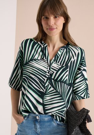 Femme portant un chemisier à manches courtes noir et blanc à motifs de feuilles tropicales avec des boutons, tenant un sac tissé noir et un jean bleu.