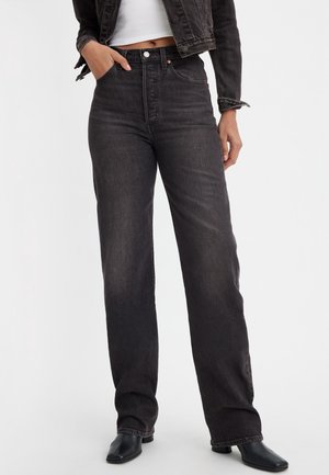 Woman wearing high-waisted, straight-leg black jeans, paired with a cropped black denim jacket, white cropped top, and black heeled shoes.