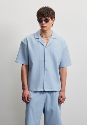 Junger Mann trägt hellblaues kurzärmeliges Hemd und passende Shorts mit dunkler Sonnenbrille und mehrlagigen Halsketten vor einfarbigem Hintergrund.