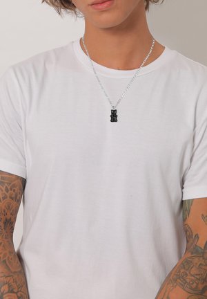 Sorthårede gummibear-vedhæng på en sølvkæde, båret med en simpel hvid t-shirt. Har fine kædelinks og synlige arm-tatoveringer.