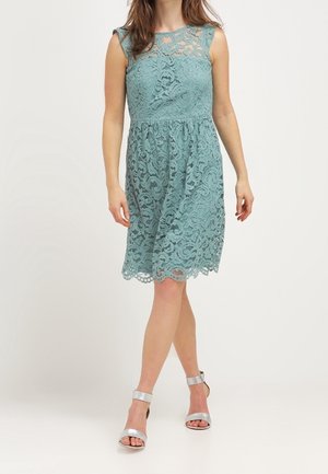 Nainen, joka pukee hihattoman polvipituisen teepukuisen pitsimekon ja hopeiset korkokengät, seisoo yksinkertaisen taustan edessä.