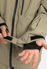 Osoba upravuje čierny suchý zips na manžete béžovej bundy s otvorom na palec, na ľavej ruke má na prste prsteň zo striebra.