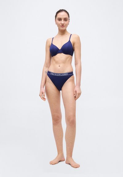 Γυναίκα που στέκεται ξυπόλητη φορώντας μπλε σουτιέν και ταιριαστά σλιπ με ζώνη μέσης "Polo Ralph Lauren" σε απλό φωτεινό φόντο.